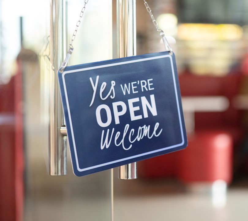 Blue sign hanging from a glass door with the words yes we're open welcome
